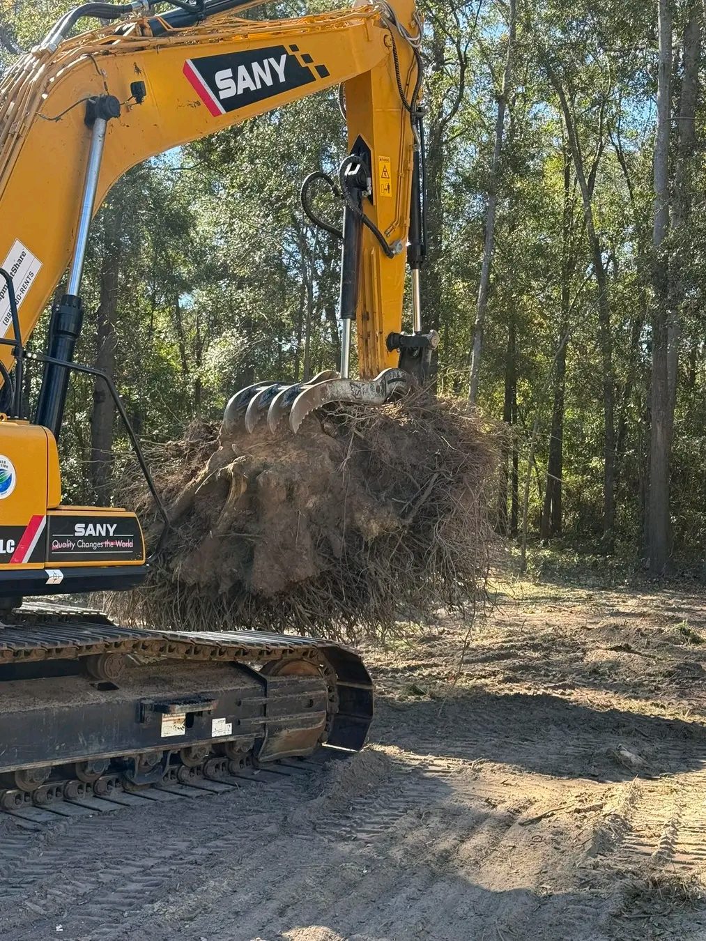 TCB Farms & Clearing stump removal project — excavator lifting a large root ball out of cleared woodland